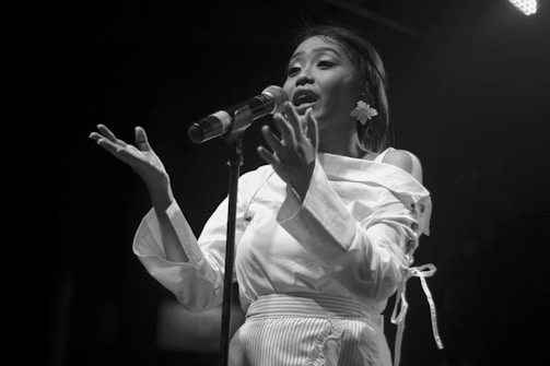 A woman passionately singing into a microphone on stage. She wears a white blouse with decorative elements and a flower in her hair. Her expressive gestures and facial expression suggest a deep emotional connection to the song.