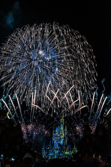 Vibrant fireworks burst in the night sky above a brightly illuminated castle, creating a spectacular and dazzling display. The colors of the fireworks reflect off the castle, enhancing its intricate towers and architecture.