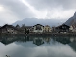 Several houses are situated by a calm lake, with their reflections visible on the water's surface. The scene is backdropped by misty mountains under a cloudy sky, creating a tranquil alpine setting.