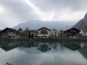 Several houses are situated by a calm lake, with their reflections visible on the water's surface. The scene is backdropped by misty mountains under a cloudy sky, creating a tranquil alpine setting.