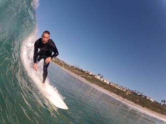 A surfer rides on a breaking wave with a look of concentration. The wave curls powerfully around him, and he is dressed in a black wetsuit. The background reveals a coastal scene with houses and palm trees lining the beach under a clear blue sky.