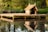 A wooden platform is situated on a tranquil pond, reflecting in the still water. Atop the platform is a small, rustic hut with a slanted roof, framed by lush greenery and wild plants along the pond's edge. Two birds are perched on the roof, adding a touch of life to the peaceful scene.