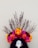 A vibrant floral headdress with a variety of colorful flowers such as orange, yellow, pink, and red, arranged in an arc. Branches extend upward from the arrangement, adding a natural, organic element. The headdress appears to be worn on a mannequin or head form with dark, long hair.