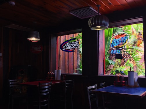 A dimly lit pub interior with wooden walls and ceiling. Two tables with chairs are visible, each set with condiments and paper towels. Neon signs in the windows display various beer brands. A window offers a view of lush green foliage outside.