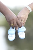 Two hands, each adorned with a ring, are holding a pair of small, blue and white baby shoes. The shoes have the words 'I love BABY' embroidered on them. The background is softly blurred, emphasizing the focus on the hands and shoes.