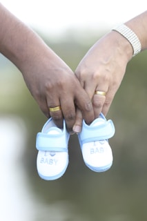 Two hands, each adorned with a ring, are holding a pair of small, blue and white baby shoes. The shoes have the words 'I love BABY' embroidered on them. The background is softly blurred, emphasizing the focus on the hands and shoes.