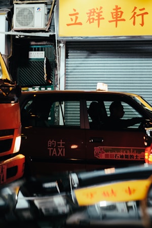 A red taxi is positioned on a city street at night with a driver inside. The taxi has Chinese characters and the word 'TAXI' written on its side. The background includes a closed shop with a yellow sign displaying Chinese text and a metallic shutter. There is also an air conditioning unit visible above the shop.