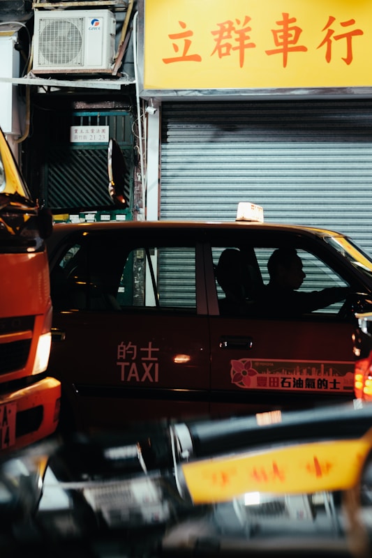 A red taxi is positioned on a city street at night with a driver inside. The taxi has Chinese characters and the word 'TAXI' written on its side. The background includes a closed shop with a yellow sign displaying Chinese text and a metallic shutter. There is also an air conditioning unit visible above the shop.