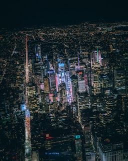 Aerial view of a bustling cityscape at night with numerous skyscrapers illuminated by bright, colorful lights. Streets are lined with streams of car lights, adding dynamic lines of red and white to the scene.