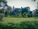 A lush green lawn is surrounded by a variety of trees and shrubs, with tall trees creating a dense backdrop. The scene captures the essence of a tranquil park setting with scattered leaves on the ground and well-maintained grass.