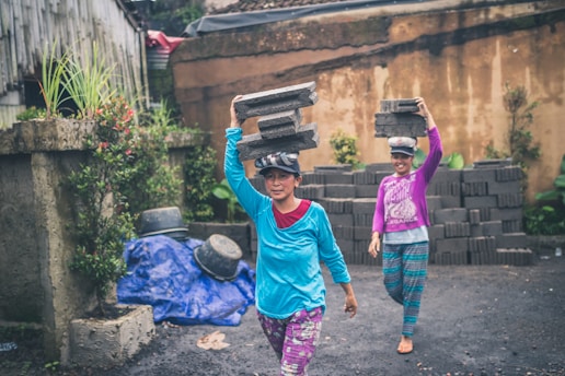 two women walking while carrying concrete bricks
