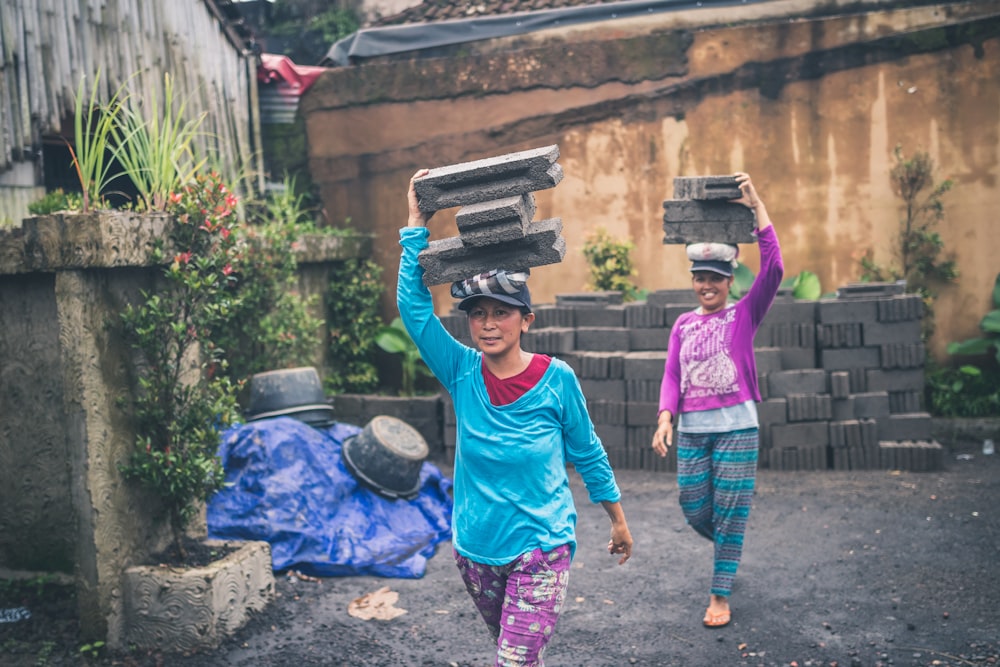 two women walking while carrying concrete bricks