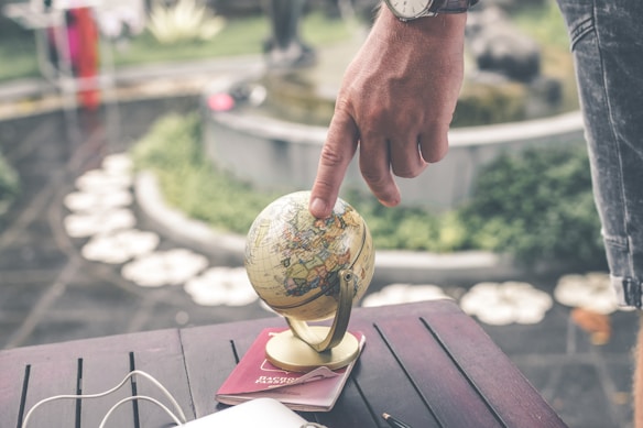 A hand with a watch is pointing at a small globe, which is resting on what appears to be a passport on a wooden table. In the background, there is greenery and a blurred view of an outdoor space, including a circular arrangement of stones or flowers.