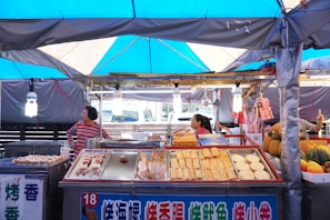 A bustling street food stall with a variety of grilled seafood and snacks on display. Two people are attending to the stand under a blue tarpaulin, surrounded by colorful produce including pineapples and melons. The atmosphere appears lively and inviting with hanging lights illuminating the scene.