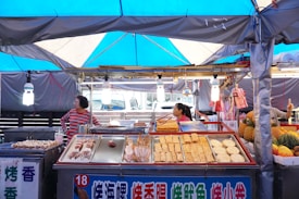 A bustling street food stall with a variety of grilled seafood and snacks on display. Two people are attending to the stand under a blue tarpaulin, surrounded by colorful produce including pineapples and melons. The atmosphere appears lively and inviting with hanging lights illuminating the scene.