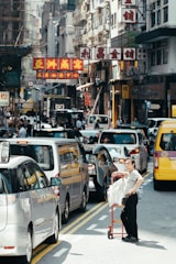 A busy urban street with a traffic jam of cars. A person stands next to a red cart loaded with packages. Multistory buildings with colorful signs featuring Chinese characters line the street. Pedestrians move along the sidewalks, and the scene is bustling with activity.