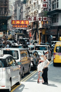 A busy urban street with a traffic jam of cars. A person stands next to a red cart loaded with packages. Multistory buildings with colorful signs featuring Chinese characters line the street. Pedestrians move along the sidewalks, and the scene is bustling with activity.