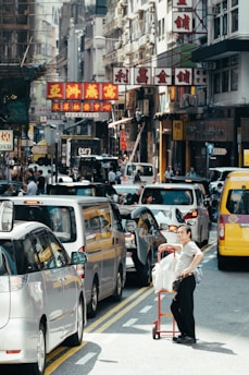 A busy urban street with a traffic jam of cars. A person stands next to a red cart loaded with packages. Multistory buildings with colorful signs featuring Chinese characters line the street. Pedestrians move along the sidewalks, and the scene is bustling with activity.
