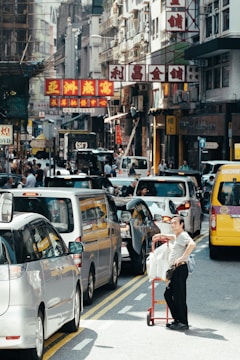 A busy urban street with a traffic jam of cars. A person stands next to a red cart loaded with packages. Multistory buildings with colorful signs featuring Chinese characters line the street. Pedestrians move along the sidewalks, and the scene is bustling with activity.
