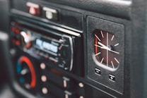 A close-up view of a car dashboard featuring an analog clock and a digital car stereo system. The clock displays time with white hands on a black background. The stereo has a digital display and several buttons, some of which are accented in red and blue.