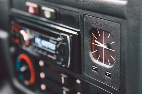 A close-up view of a car dashboard featuring an analog clock and a digital car stereo system. The clock displays time with white hands on a black background. The stereo has a digital display and several buttons, some of which are accented in red and blue.