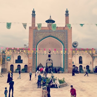 A large courtyard features an ornate building with detailed tile work and two tall minarets. The architecture has arches and intricate patterns, typical of Islamic design. Various people are walking, sitting, and socializing, some wearing traditional clothing. Colorful flags are strung across the courtyard above the people, adding a festive feel.