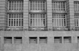 A grayscale image of a building facade featuring a series of large, grid-like windows separated by vertical stone columns. Below the windows, several security cameras are mounted on a concrete wall with rectangular air vents.