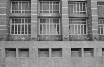 A grayscale image of a building facade featuring a series of large, grid-like windows separated by vertical stone columns. Below the windows, several security cameras are mounted on a concrete wall with rectangular air vents.