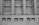 A grayscale image of a building facade featuring a series of large, grid-like windows separated by vertical stone columns. Below the windows, several security cameras are mounted on a concrete wall with rectangular air vents.