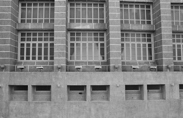 A grayscale image of a building facade featuring a series of large, grid-like windows separated by vertical stone columns. Below the windows, several security cameras are mounted on a concrete wall with rectangular air vents.