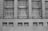 A grayscale image of a building facade featuring a series of large, grid-like windows separated by vertical stone columns. Below the windows, several security cameras are mounted on a concrete wall with rectangular air vents.