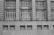 A grayscale image of a building facade featuring a series of large, grid-like windows separated by vertical stone columns. Below the windows, several security cameras are mounted on a concrete wall with rectangular air vents.