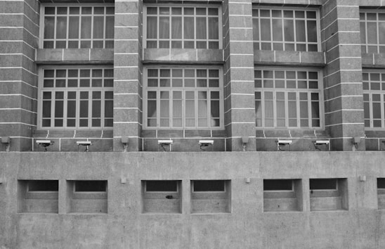A grayscale image of a building facade featuring a series of large, grid-like windows separated by vertical stone columns. Below the windows, several security cameras are mounted on a concrete wall with rectangular air vents.