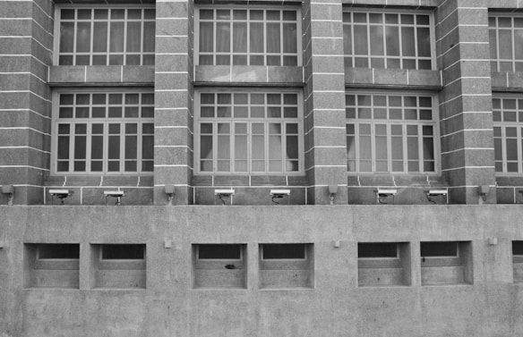 A grayscale image of a building facade featuring a series of large, grid-like windows separated by vertical stone columns. Below the windows, several security cameras are mounted on a concrete wall with rectangular air vents.