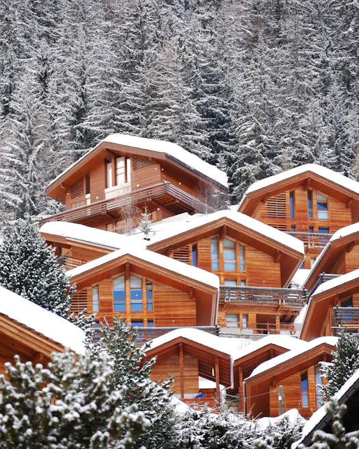 Wooden chalets with triangular roofs are nestled on a snowy hillside, surrounded by a dense forest of snow-dusted evergreen trees.