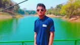 A person is standing on a bridge or walkway, wearing sunglasses and a navy blue polo shirt with the letters 'CISC' on it. The background features a vibrant, green-colored river or lake with trees on both sides, some of which are lush and others are more sparse. The setting appears to be peaceful and surrounded by nature.