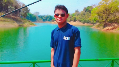 A person is standing on a bridge or walkway, wearing sunglasses and a navy blue polo shirt with the letters 'CISC' on it. The background features a vibrant, green-colored river or lake with trees on both sides, some of which are lush and others are more sparse. The setting appears to be peaceful and surrounded by nature.