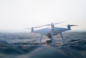 A drone rests on a sandy beach with a soft, hazy sky in the background, capturing a serene and peaceful atmosphere. The lighting is gentle, emphasizing the smooth texture of the sand and the sleek design of the drone.