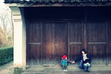 A young child in a red hat and colorful jacket sits on a stone step in front of a large wooden door, holding hands with an adult who is seated nearby. The adult is wearing a dark jacket with a light scarf, and they are smiling at each other. The architecture features dark wooden beams and a white column. In the background, there are trees and a stone path leading to the door.