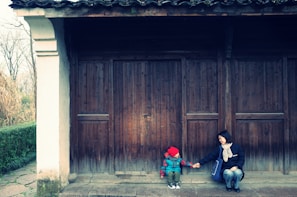 A young child in a red hat and colorful jacket sits on a stone step in front of a large wooden door, holding hands with an adult who is seated nearby. The adult is wearing a dark jacket with a light scarf, and they are smiling at each other. The architecture features dark wooden beams and a white column. In the background, there are trees and a stone path leading to the door.