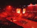 An indoor ice hockey arena filled with a large crowd, bathed in red lighting. A giant central scoreboard projects fiery visuals, illuminating the ice and audience. The atmosphere is charged, likely during a live sporting event.