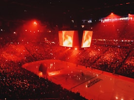 An indoor ice hockey arena filled with a large crowd, bathed in red lighting. A giant central scoreboard projects fiery visuals, illuminating the ice and audience. The atmosphere is charged, likely during a live sporting event.