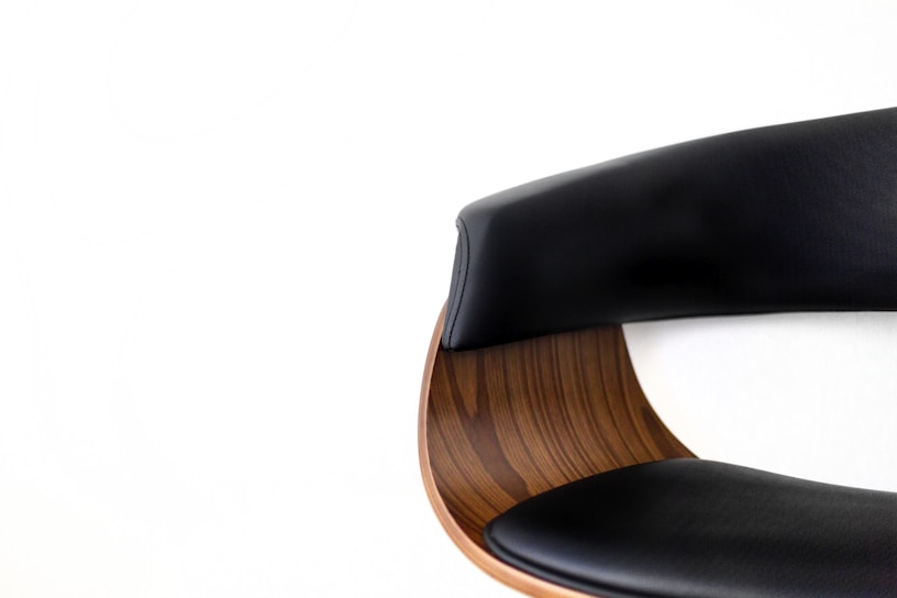 A close-up view of a modern chair featuring a curved wooden backrest and upholstered black leather cushions. The sleek design combines natural wood grain texture with smooth, dark leather, creating a blend of contemporary and classic styles against a plain white background.
