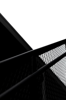 A black metal fence with a diamond-shaped pattern is captured against a contrasting bright white sky. A dark, angular structure is partially visible, creating a stark geometric contrast in the composition.