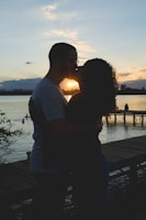 A silhouetted couple is kissing against the backdrop of a sunset by a lake. In the background, there is a wooden dock extending into the water and other people swimming. The scene is calm and romantic, with trees partially framing the image.
