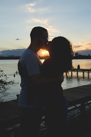 A silhouetted couple is kissing against the backdrop of a sunset by a lake. In the background, there is a wooden dock extending into the water and other people swimming. The scene is calm and romantic, with trees partially framing the image.