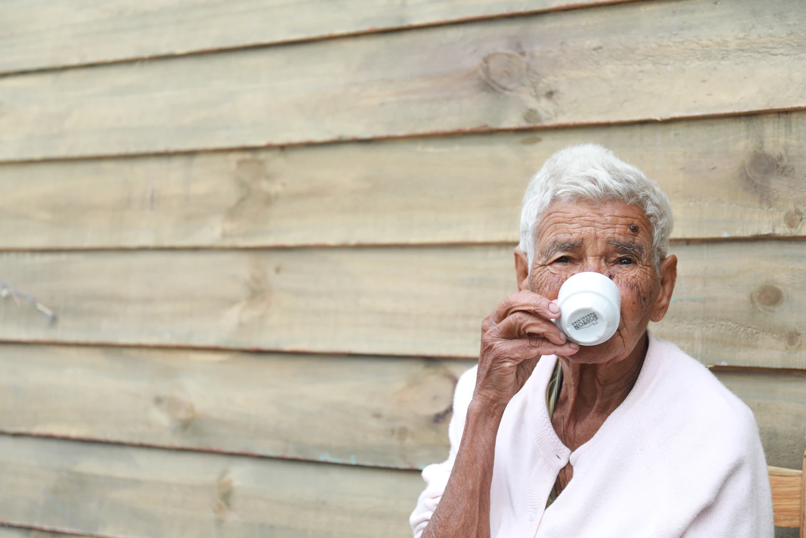 An older woman with white hair holding a teacup close to her face, her hands and expression showing years of lived experience