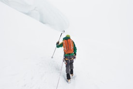 A lone climber is trekking through a snowy landscape. The climber wears a bright green jacket and an orange backpack, using a pole to navigate the snow. The scene is dominated by white snow and a foggy sky, creating an isolated and challenging environment.
