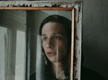 A person looks through an old, weathered mirror with a distressed wooden frame. The mood appears introspective and somber, with soft lighting casting gentle shadows.