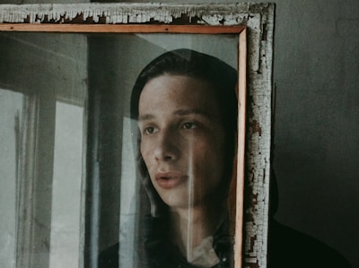 A person looks through an old, weathered mirror with a distressed wooden frame. The mood appears introspective and somber, with soft lighting casting gentle shadows.